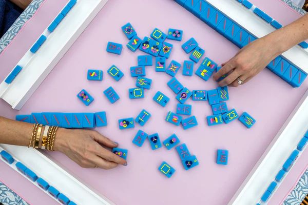 Hands playing with bright blue custom mahjong tiles on a pink table